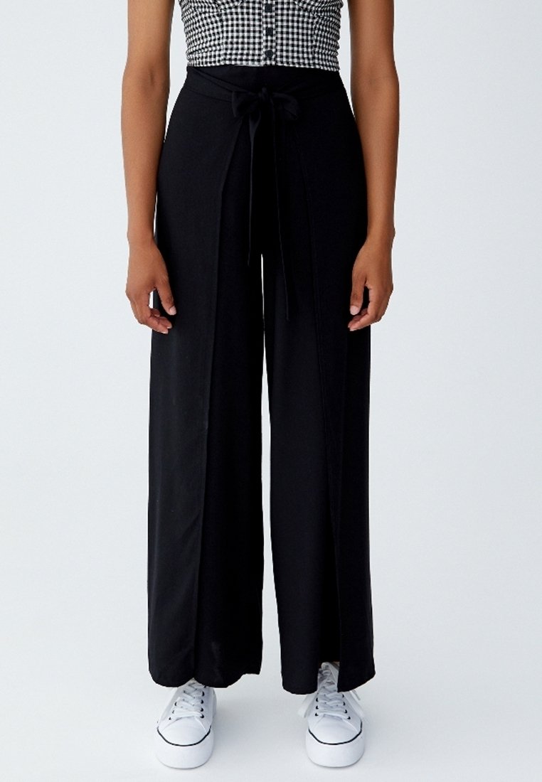 Zwarte wijde broek met een strikband, soepel materiaal en voorzakken. Gecombineerd met witte sneakers, wat een casual stijl laat zien.