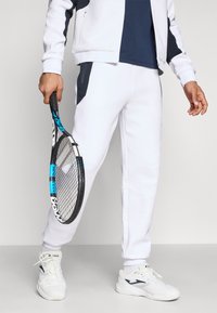 Biały dres sportowy z granatowymi akcentami, z suwakiem-kieszoniem, zestawiony z białymi trampkami. Osoba trzyma rakietę tenisową.