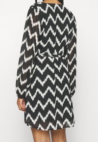 Svart och vit klänning med vågigt mönster, långa ärmar, insvängd midja och lös passform, tillverkad av lätt, genomskinlig tyg.