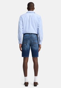 Blå denimshorts med lige snit, der har to baglommer og stikninger. Kombineret med en lyseblå skjorte med knapper.