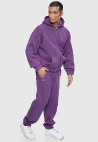Lila Kapuzenpulli und passende Jogginghose, hergestellt aus weichem Material. Der Kapuzenpulli hat einen Reißverschluss und Taschen; die Hose verfügt über einen Kordelzugbund und eine Reißverschlusstasche.