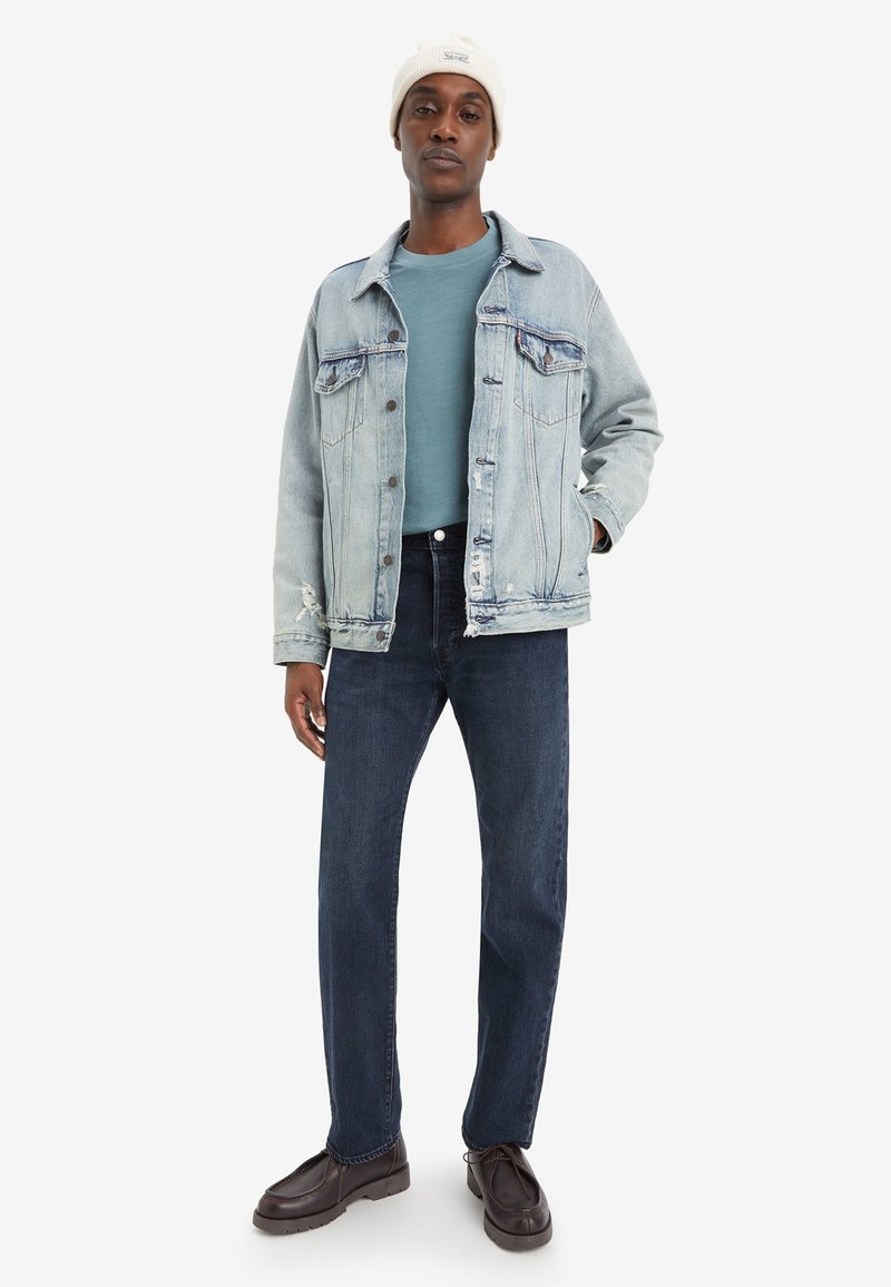 Lichtblauwe spijkerjack over een teal trui, gecombineerd met donkere spijkerbroek en bruine schoenen. De look wordt gecompleteerd met een crèmekleurige gebreide beanie.