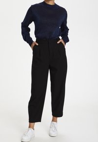 Persoon in een marineblauwe longsleeve top, zwarte hoge broek met handen in de zakken, en witte sneakers op een witte achtergrond.