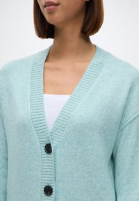 Ανοιχτό μπλε πλεχτό καρέ νάνο με V-λαιμόκοψη, κλείσιμο με κουμπιά και ριμπ μανσέτες, με μαλακή υφή και ελαφρώς χνουδωτή εμφάνιση.