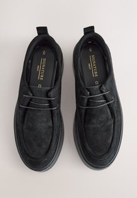 Sorte ruskindssko med rundet tå, slip-on design, med et dekorativt snørebånd og en tekstureret gummisål. Minimalistisk branding synlig.