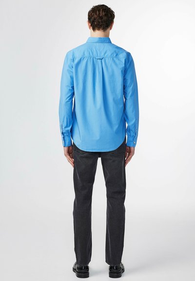 Lichtblauwe longsleeve shirt met een kraag, knoopmanchetten en een gebogen zoom. Gekozen bij donkere jeans en zwarte schoenen, gezien vanaf de achterkant.