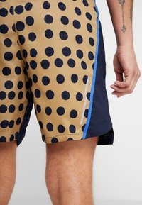 Short beige avec de gros pois noirs et une bande bleue sur le côté, porté par une personne avec un bras tatoué.