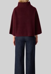 Woman wearing a ribbed burgundy turtleneck sweater with wide sleeves and dark blue wide-legged pants, standing facing away.