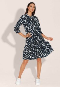 Navy-Blaues Kleid mit weißen Punkten, kragenförmiges Design, Dreiviertelärmel und Rüschensaum. Kombiniert mit weißen Sneakers.