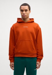 Felpa arancione con tasca frontale e cappuccio regolabile con cordino. Presenta il logo bianco "Calvin Klein" sul petto. Tessuto morbido al tatto.