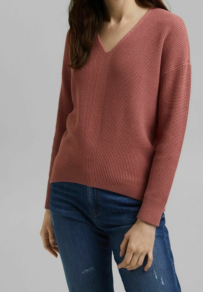 Pull en maille côtelée, de couleur rose, avec un col en V et une coupe décontractée. Manches longues et motif texturé. Associé à un jeans en denim bleu.