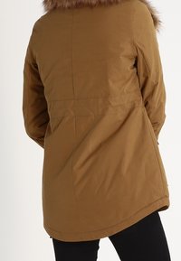 Brun parkasjacka med avtagbar fuskpäls, utrustad med midjesnörning och rundad fåll, tillverkad av ett slätt, slitstarkt tyg.