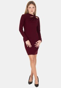 Vestito bordeaux a maniche lunghe con dettaglio annodato al collo; silhouette aderente e lunghezza al ginocchio; tessuto liscio, leggermente strutturato.