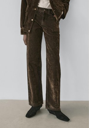 Persona che indossa pantaloni di velluto marrone scuro, stivali neri con punta a punta e una giacca di velluto abbinata, con una mano in tasca.