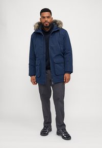 Parka impermeable azul marino con capucha forrada de piel, dos bolsillos frontales y cierre de cremallera, combinada con pantalones grises y zapatillas deportivas negras.