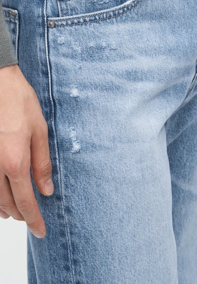 Αριστερό χέρι που ξεκουράζεται κοντά στην μπροστινή τσέπη ενός ανοιχτόχρωμου τζιν με φθορές, με ορατά ραφές και υφή υφάσματος.