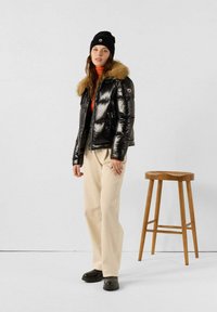 Schwarze Pufferjacke mit Pelzkragen, orangefarbener Rollkragenpullover, cremefarbene Hose, schwarze Mütze und Clogs; steht neben einem Holzschemel.