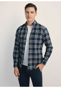 Granatowa koszula flanelowa w kratę z białymi i beżowymi paskami, z kołnierzykiem na guziki i jedną kieszenią na piersi, noszona na białej koszulce.