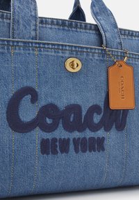 Sac fourre-tout en denim avec "Coach New York" brodé en bleu foncé et une étiquette en cuir marron Coach suspendue à une chaîne à billes dorée.