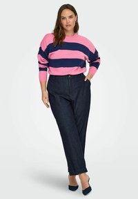 Kobieta stoi z jedną ręką w kieszeni, ubrana w różowo-granatowy sweter w paski z długim rękawem, ciemnoniebieskie spodnie i czarne, spiczaste buty.