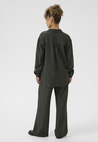 Donna con capelli ricci sta in piedi di spalle, indossando una camicia oversize a righe scure e pantaloni larghi coordinati, insieme a scarpe nere.