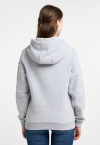 Jonge vrouw met bruin haar in een staart, gekleed in een effen lichtgrijze hoodie en blauwe spijkerbroek, gezien van achteren tegen een witte achtergrond.