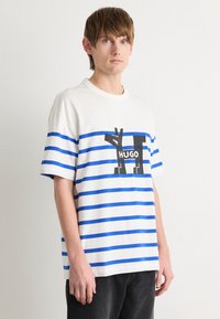 Weißes T-Shirt mit blauen horizontalen Streifen, auf der Vorderseite mit einem großen schwarzen Grafik und dem Text "HUGO" gestaltet. Lockerer Schnitt und kurze Ärmel.