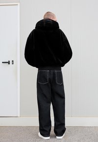 Persona in piedi con le spalle rivolte, che indossa una giacca di pelliccia nera, jeans larghi scuri con cuciture bianche e sneakers bianche, contro un muro semplice.