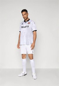 Egy férfi áll fehér és világosszürke focimezben, amelyen a "TRUST IT" felirat olvasható, fekete galléros ingben, fehér nadrágban, zokniban és stoplis cipőben, egyszínű háttér előtt.