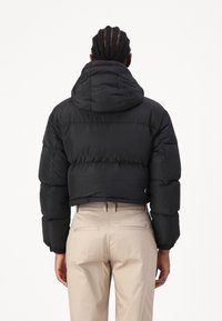 Persona con capelli intrecciati che indossa una giacca a vento corta nera e pantaloni beige, vista da dietro su uno sfondo bianco.