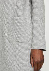 Cappotto grigio testurizzato con una tasca quadrata visibile, colletto semplice e maniche con risvolto, che mostra un dettaglio in tessuto intrecciato.