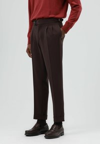 Bruine geplooide broek met een getailleerde pasvorm, voorzien van zijriempjes, een gladde textuur, omgeslagen zoom en gedragen met bruine loafers.