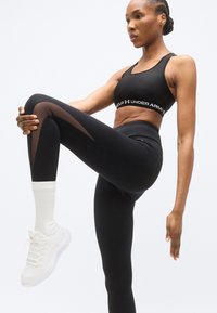 Μαύρο αθλητικό σουτιέν με λογότυπο, μαύρα κολάν με τμήματα από δίχτυ, και λευκά αθλητικά παπούτσια. Λείο ύφασμα, εφαρμοστή γραμμή με ψηλή μέση.