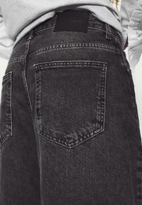 Gros plan sur les poches arrière d'un jean en denim noir porté avec une chemise gris clair rentrée à l'intérieur.