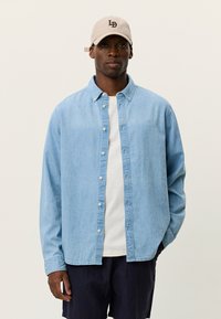 Ανοιχτό μπλε πουκάμισο από τζιν με κουμπιά, γιακά, μακριά μανίκια και λευκά κουμπιά, φορεμένο πάνω από λευκό T-shirt και συνδυασμένο με σκούρα σορτς.