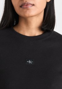 Zwarte geribbelde sweatshirt met een ronde halslijn en een klein rechthoekig Calvin Klein Jeans-logotag op de borst.