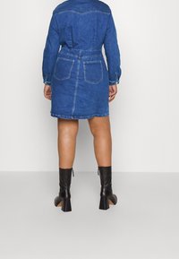 Vestido de mezclilla en azul medio con puños abotonados, cintura ceñida con cinturón y dos bolsillos traseros. Combinado con botas negras de tacón alto hasta la rodilla.