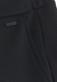 Gros plan sur un tissu noir avec une poche et une manche côtelée, avec un petit logo rectangulaire noir embossé "4F".