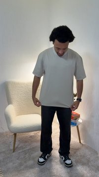 Light beige, short-sleeved t-shirt with a crew neckline, made of smooth fabric. Paired with dark pants and black sneakers, standing indoors.