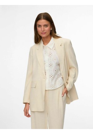Femme portant un costume crème avec un pantalon à jambes larges et un blazer assorti sur un chemisier blanc à motif ajouré, debout devant un fond blanc.