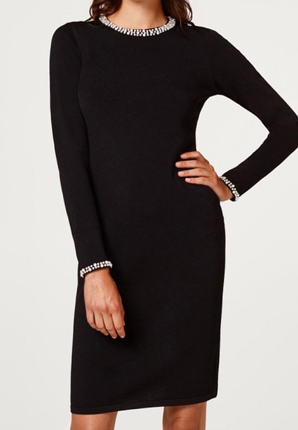 Robe noire mi-longue à manches longues avec bordure perlée blanche sur l'encolure et les poignets, portée par une personne avec une main sur la hanche et l'autre détendue.