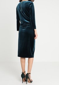 Sammetsteal klänning med draperad rygg och långa ärmar, med en dold dragkedja. Kombinerad med svarta högklackade sandaler.