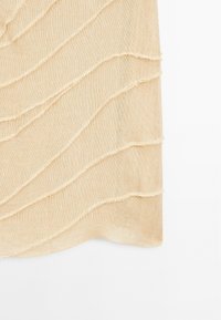 Tissu beige avec un motif de couture ondulée et des bords effilochés, créant un aspect texturé. Matériau léger et transparent.