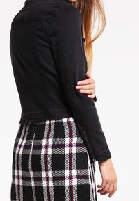 Chaqueta de mezclilla negra con un diseño ajustado, con un largo recortado, cierre de botones en la parte delantera y cuello estándar. Llevada sobre una falda de cuadros.