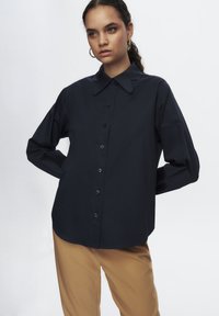 Blusa negra de manga larga con cuello clásico, cierre de botones y corte relajado. Textura suave, mangas amplias y diseño sutil.
