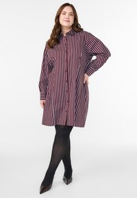 Sukienka koszulowa w bordowe i białe pionowe paski. Posiada kołnierzyk, długie rękawy i boczne kieszenie, zestawiona z czarnymi rajstopami i szpilkami.