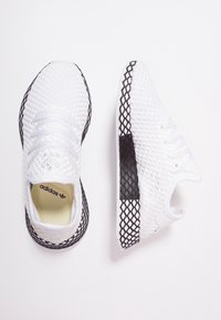 Vita nätliknande sportskor i vitt med en texturerad yta, svarta accenter och en kontrasterande nätliknande sula. Har snören och varumärkeslogotyp inuti.