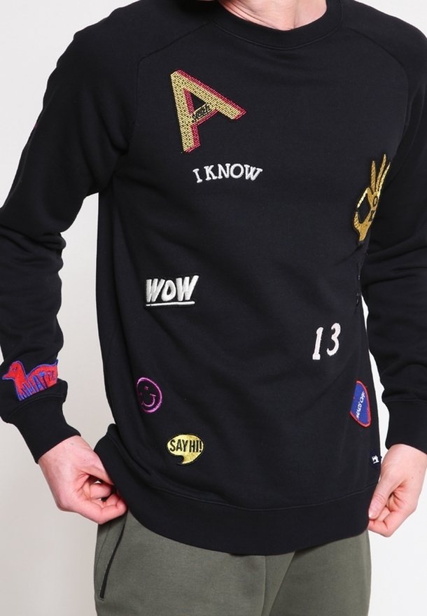 Sweat-shirt noir avec des patchs brodés colorés comprenant "I KNOW", "WOW", "13", un visage souriant et "SAY HI!" porté par une personne.