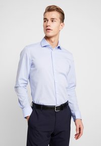 Camicia azzurra a maniche lunghe con colletto alla francese, indossata con pantaloni scuri e una cintura nera. Tessuto liscio e vestibilità su misura.
