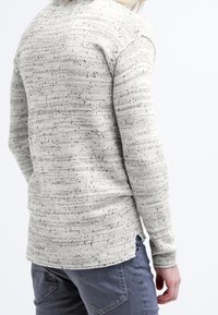 Ljust grå stickad tröja med en strukturerad, ojämn mönstring, långa ärmar och en något krökt nederkant. Bar med grå byxor, sedd bakifrån.